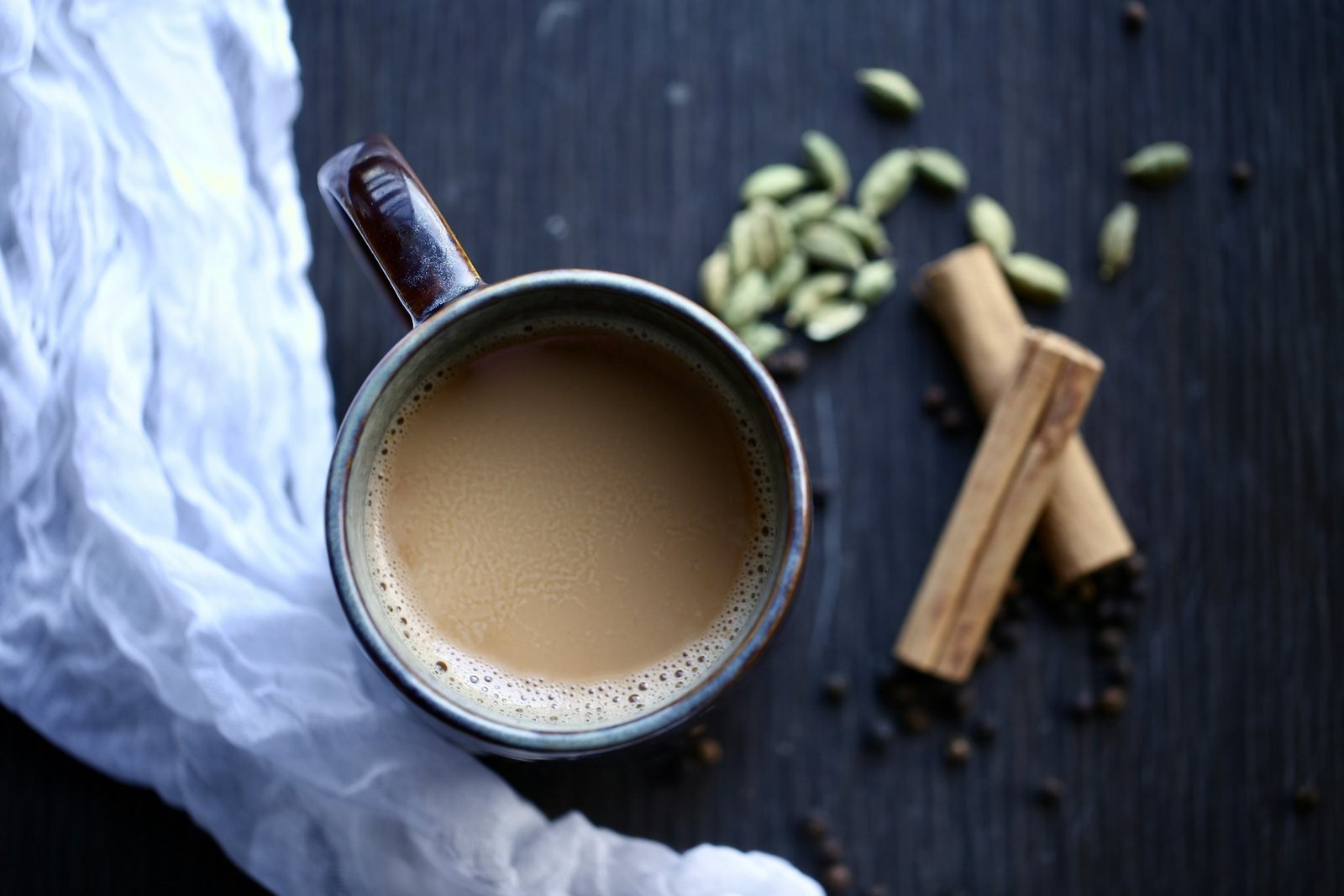 A mug of milky chai with green cardamom pods and cinnamon on a dark table